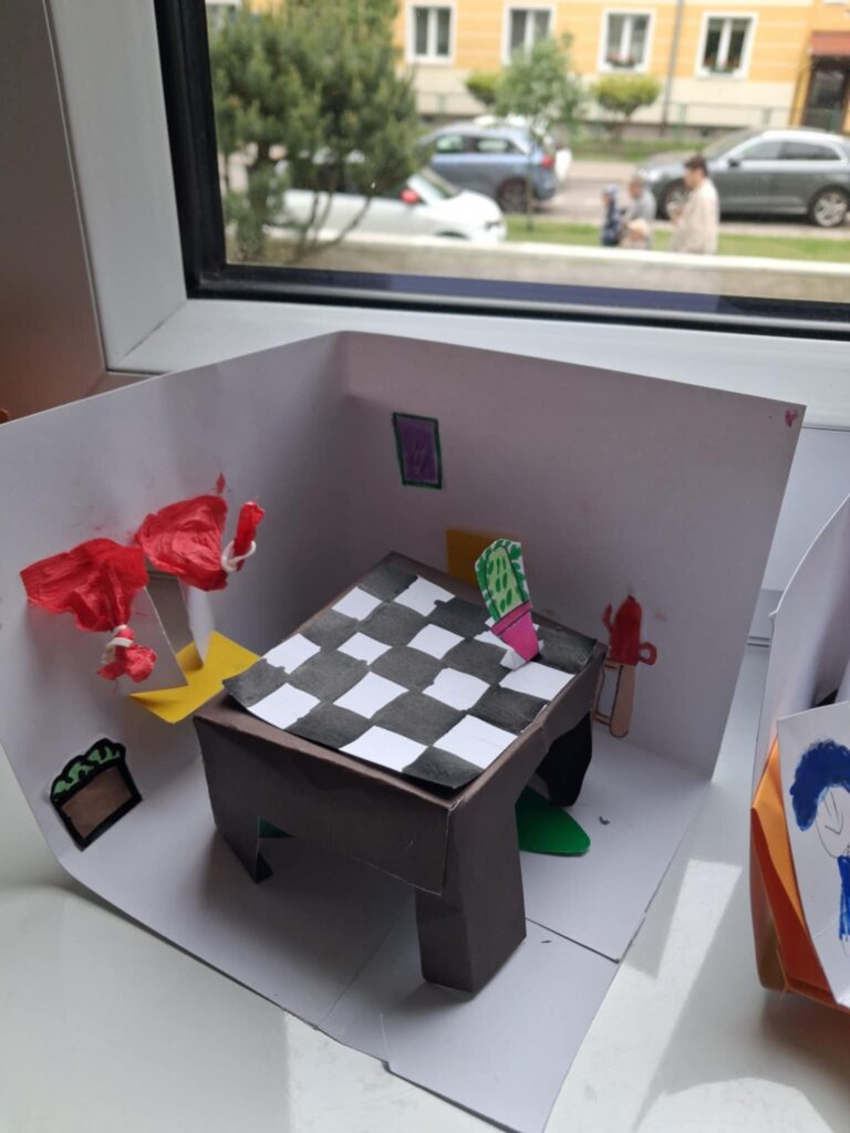 Fotografia przedstawia makietę biura detektywistycznego, wykonaną z papieru. Jest stolik i krzesełka. Wykonane sa okna z firankami.