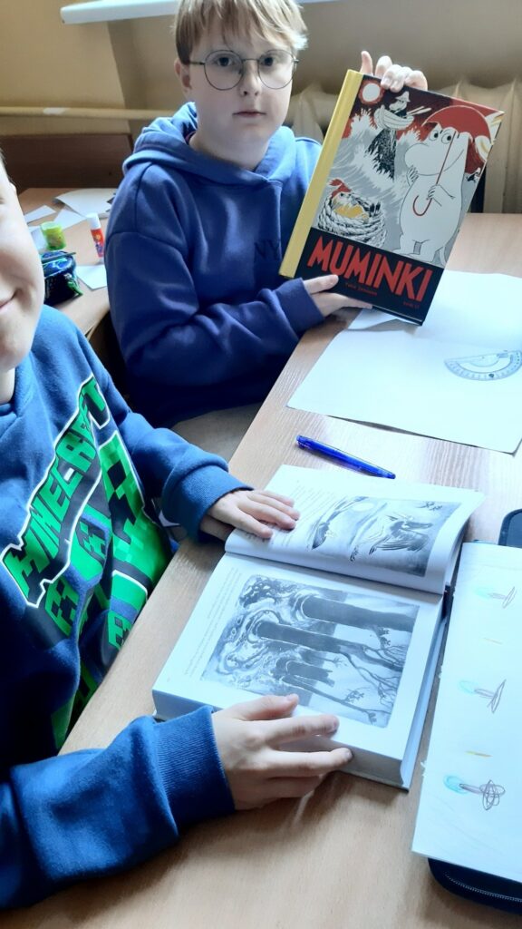 Zdjęcie przedstawia dwóch chłopców siedzących przy ławce szkolnej. Jeden z nich ustawiony przodem do fotografa trzyma w dłoniach książkę na której okładce znajduje się tytuł Muminki oraz grafika Muminków. Chłopiec ma okulary niebieską bluzę. Przed chłopcem leży kartka na kartce cyrkiel ekierka. Obok siedzi drugi chłopiec którego widać tylko niewielki fragment twarzy oraz ręce ułożone na książce która leży przed nim. Chłopiec ma niebieską bluzę z zielonym napisem.