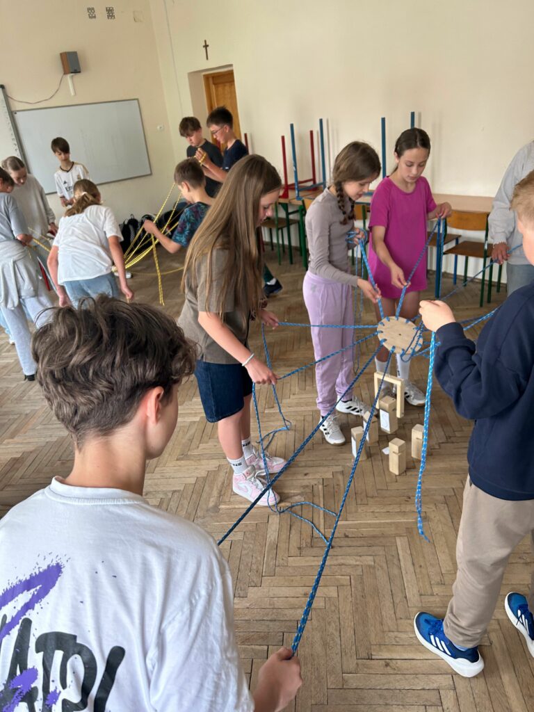 Na zdjęciu widzimy dwie grupy dzieci stojących w kręgu w sali lekcyjnej. Każde z nich trzyma w rękach sznurek, który jest przymocowany do wspólnego mechanizmu umożliwiającego przenoszenie klocków. Dzieci wspólnie budują wieżę z drewnianych elementów, biorą udział w zabawie rozwijającej współpracę. W tle widoczne są meble szkolne, prace plastyczne na ścianach oraz naturalne światło wpadające przez okna. Atmosfera na zdjęciu wydaje się być skupiona i zaangażowana.