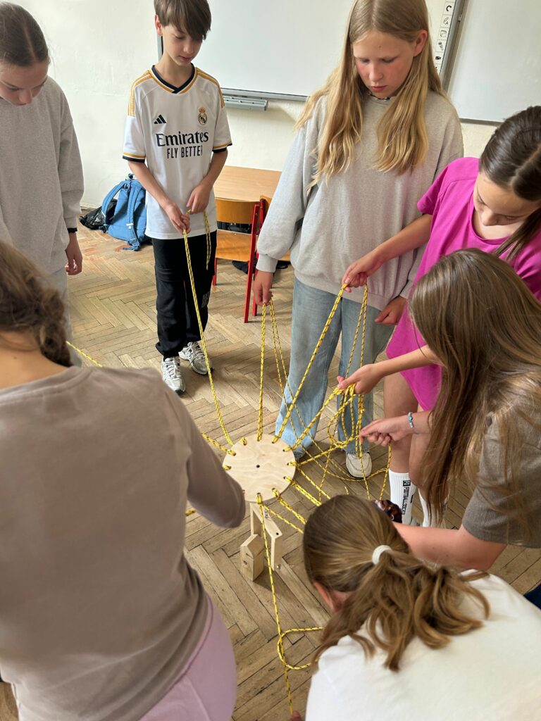 Na zdjęciu widzimy grupę dzieci stojących w kręgu w sali lekcyjnej. Każde z nich trzyma w rękach sznurek, który jest przymocowany do wspólnego mechanizmu umożliwiającego przenoszenie klocków. Dzieci wspólnie budują wieżę z drewnianych elementów, biorą udział w zabawie rozwijającej współpracę. W tle widoczne są meble szkolne, prace plastyczne na ścianach oraz naturalne światło wpadające przez okna. Atmosfera na zdjęciu wydaje się być skupiona i zaangażowana.