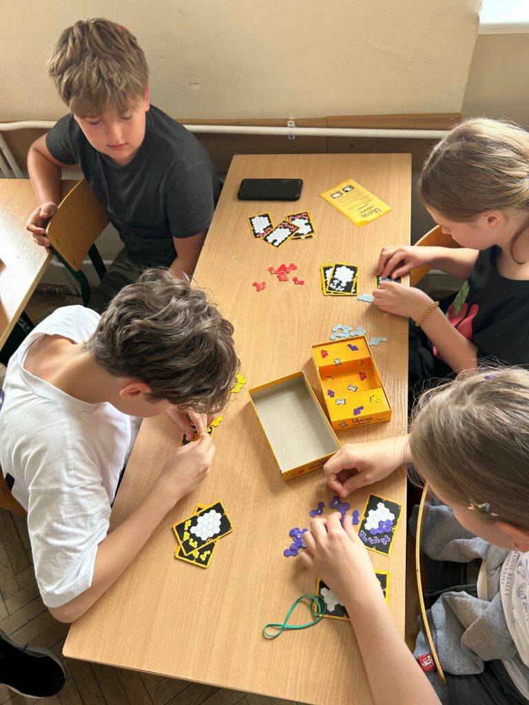 Na zdjęciu widzimy czworo dzieci siedzących przy szkolnej ławce i grających w grę planszową. Skupiają się na układaniu kolorowych elementów na kartach, prawdopodobnie rozwiązując logiczne zadania. Na stole leży pudełko od gry oraz instrukcja. Dzieci wyglądają na zaangażowane i skoncentrowane, co sugeruje, że gra wymaga myślenia i planowania. W tle widać typowe wyposażenie klasy – krzesła, ławki oraz ścianę z grzejnikiem.