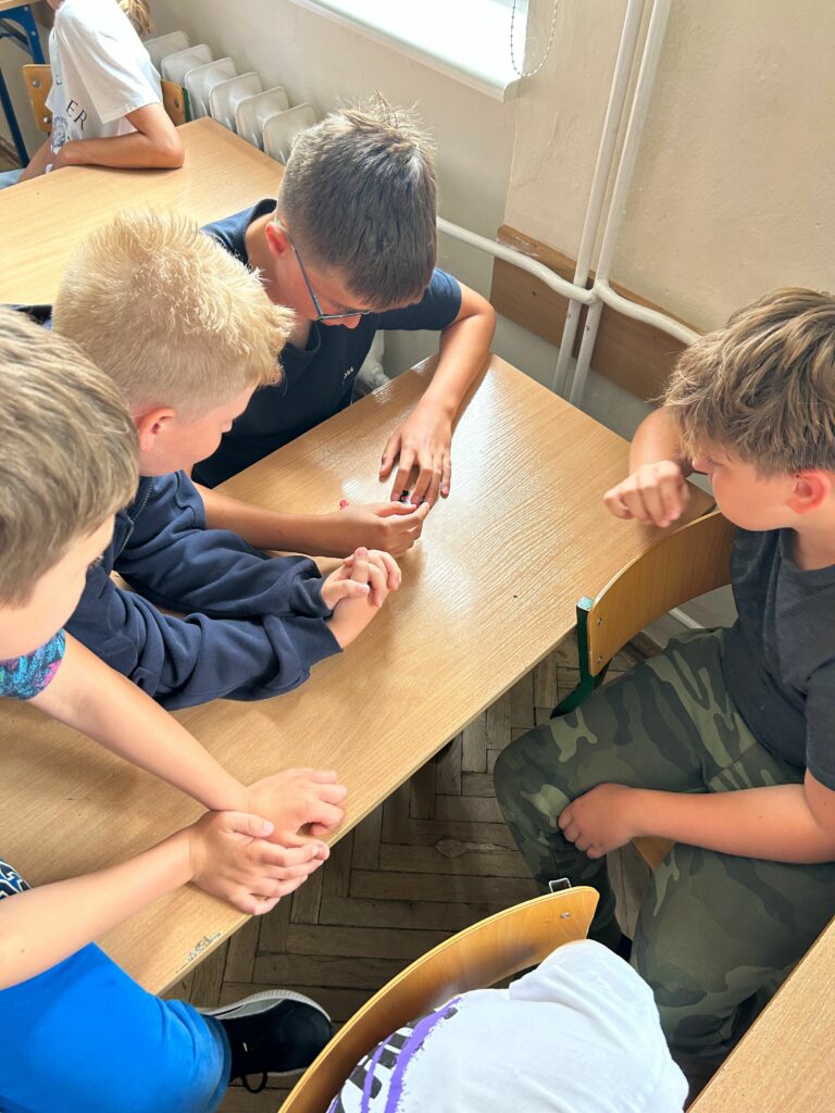 Na zdjęciu widzimy grupę chłopców siedzących przy szkolnej ławce, uważnie obserwujących coś na jej powierzchni. W centrum ich uwagi znajduje się gra/puzzle logiczne – prawdopodobnie kostka do gry. Chłopcy wydają się być skupieni i zaangażowani w rozgrywkę lub zabawę wymagającą zręczności albo szczęścia. W tle widoczny jest fragment sali lekcyjnej z grzejnikiem i oknem. Atmosfera zdjęcia sugeruje koleżeńską współpracę i wspólne spędzanie czasu.