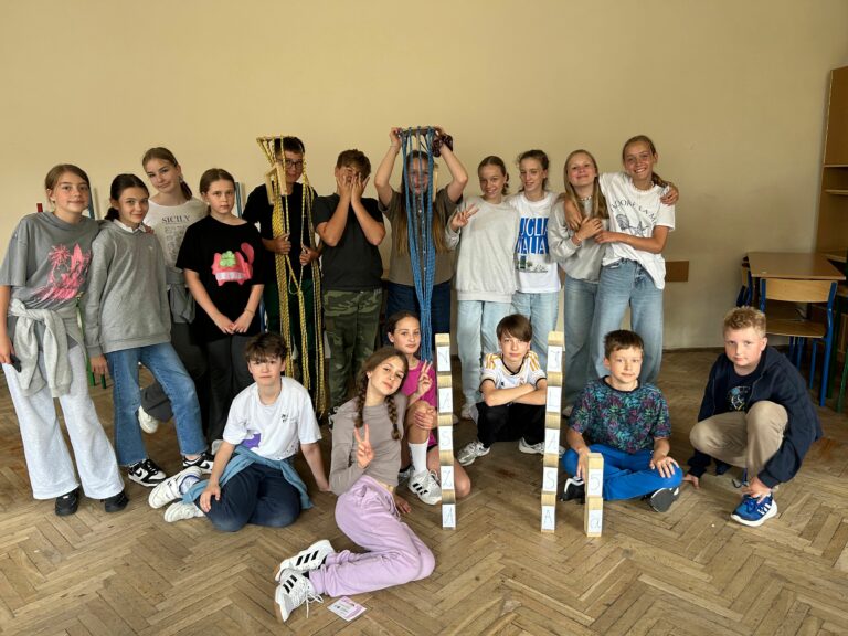 W sali lekcyjnej grupa dzieci w dwóch rzędach - kucające a przed nimi dwie wieże z kloców z napisem Nasza Klasa 5a; w drugim rzędzie dzieci stoją uśmiechnięte.