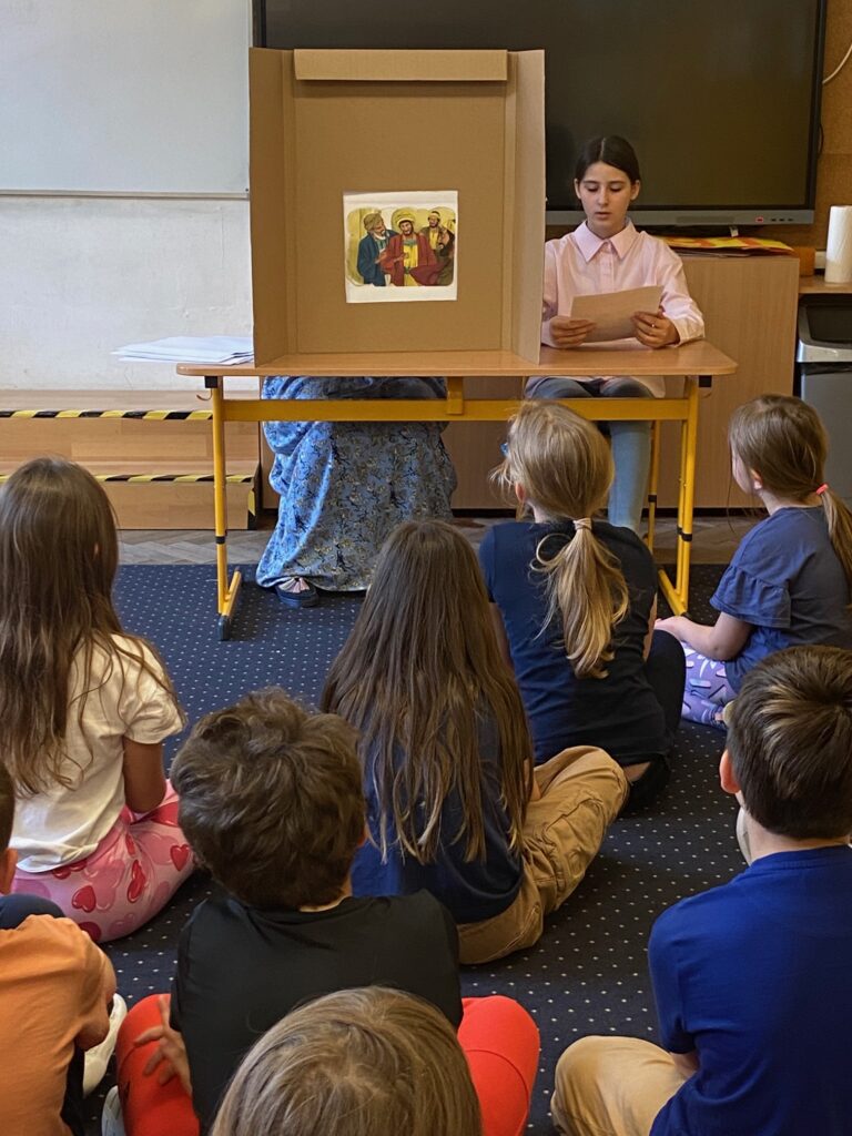 Zdjęcie przedstwia dziewczynkę siedzącą za stołem na którym umieszczono teatrzyk. Dziewczynka ma w dłoniach kartkę. Przed teatyrzykiem na podłodze tyłem do fotografa siedzą dzieci oglądające wystąpienie.