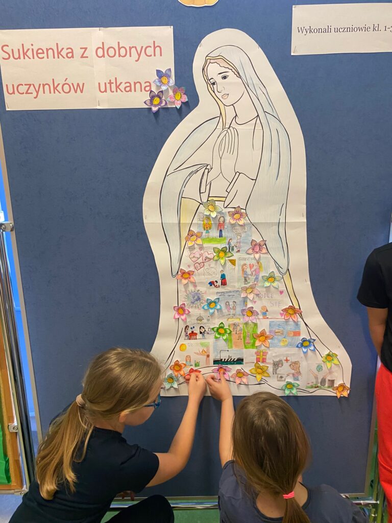 Zdjęcie przedstawia dzieci które przyczepiają na sukni Maryi kolorowe kwiart. Figura Maryi jest biało czarna przyczepiona na niebieskiej tablicy