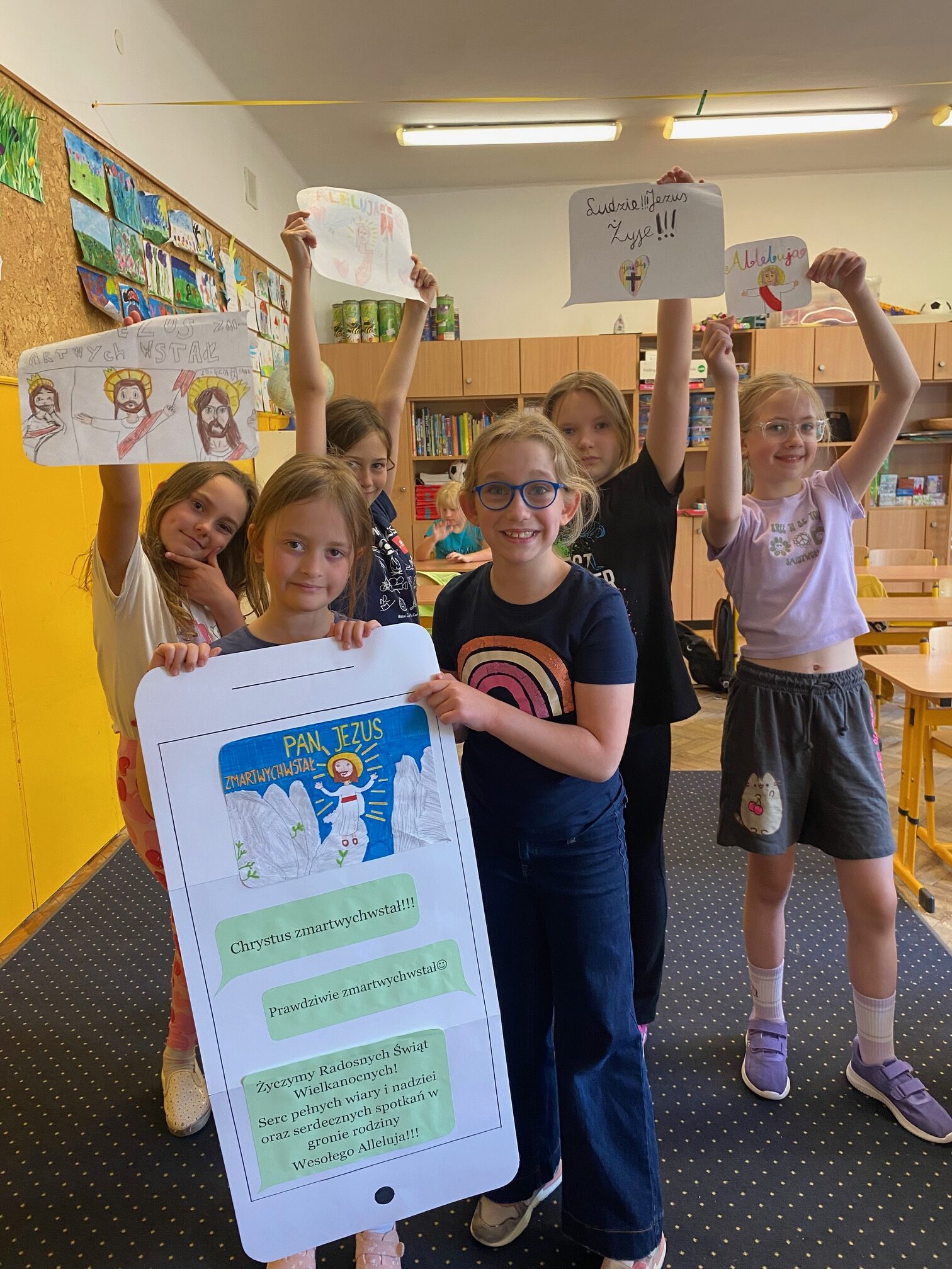 Zdjęcie przedstawia dzieci stojące w sali lekcyjnej. Trzymajace prace plastyczne przedstawiające z martwych wystałego Jezusa