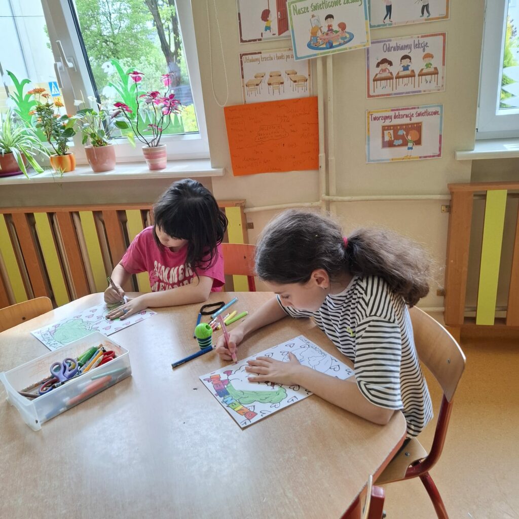 Na zdjęciu widać dwie dziewczynki siedzące przy okrągłym stole i kolorujące obrazki. Na stole leżą różnokolorowe przybory plastyczne, takie jak flamastry, kredki i nożyczki. W tle znajdują się okna z doniczkami pełnymi roślin oraz kolorowe plakaty z zasadami świetlicy. Pomieszczenie wygląda na jasne, przyjazne i zachęcające do kreatywnej zabawy.