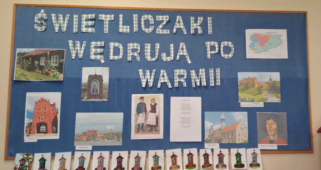 Zdjęcie przedstawia kolorową tablicę edukacyjną o tematyce regionalnej pt. "Świetliczaki wędrują po Warmii". Znajdują się na niej zdjęcia charakterystycznych miejsc Warmii, takich jak Olsztyn, Frombork i Barczewo, oraz ilustracje warmińskich kapliczek. W centralnej części widnieje para w tradycyjnych strojach ludowych i tekst hymnu Warmii. W prawym dolnym rogu znajduje się portret Mikołaja Kopernika, związanego z tym regionem. Całość ozdobiona jest dziecięcymi pracami, co wskazuje na zaangażowanie uczniów w poznawanie kultury i historii Warmii.