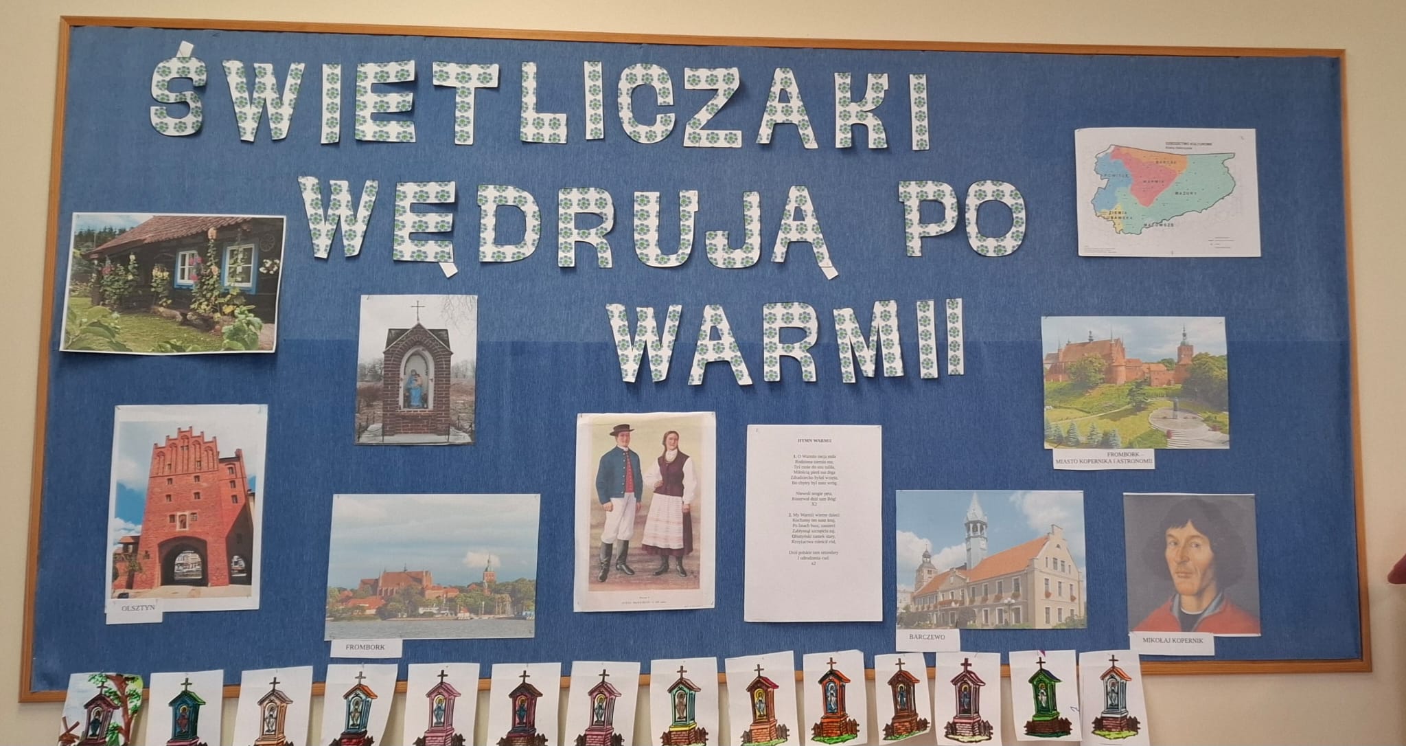 Zdjęcie przedstawia kolorową tablicę edukacyjną o tematyce regionalnej pt. "Świetliczaki wędrują po Warmii". Znajdują się na niej zdjęcia charakterystycznych miejsc Warmii, takich jak Olsztyn, Frombork i Barczewo, oraz ilustracje warmińskich kapliczek. W centralnej części widnieje para w tradycyjnych strojach ludowych i tekst hymnu Warmii. W prawym dolnym rogu znajduje się portret Mikołaja Kopernika, związanego z tym regionem. Całość ozdobiona jest dziecięcymi pracami, co wskazuje na zaangażowanie uczniów w poznawanie kultury i historii Warmii.