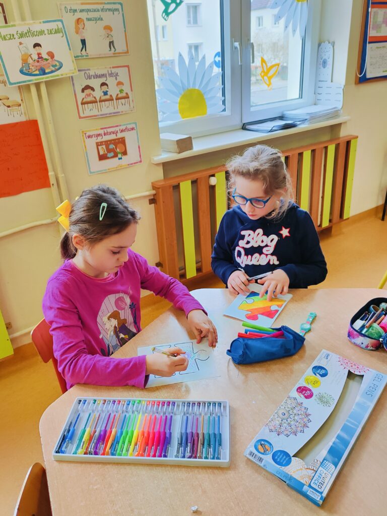Dwie dziewczynki siedzą przy stoliku w klasie i kolorują duże elementy puzzli. Na stole leżą kolorowe pisaki, piórniki i opakowanie z zestawem żelowych długopisów. W tle widać okno z dekoracjami wiosennymi oraz plakaty z zasadami zachowania. Dziewczynki skupione są na pracy, co tworzy spokojną i twórczą atmosferę.