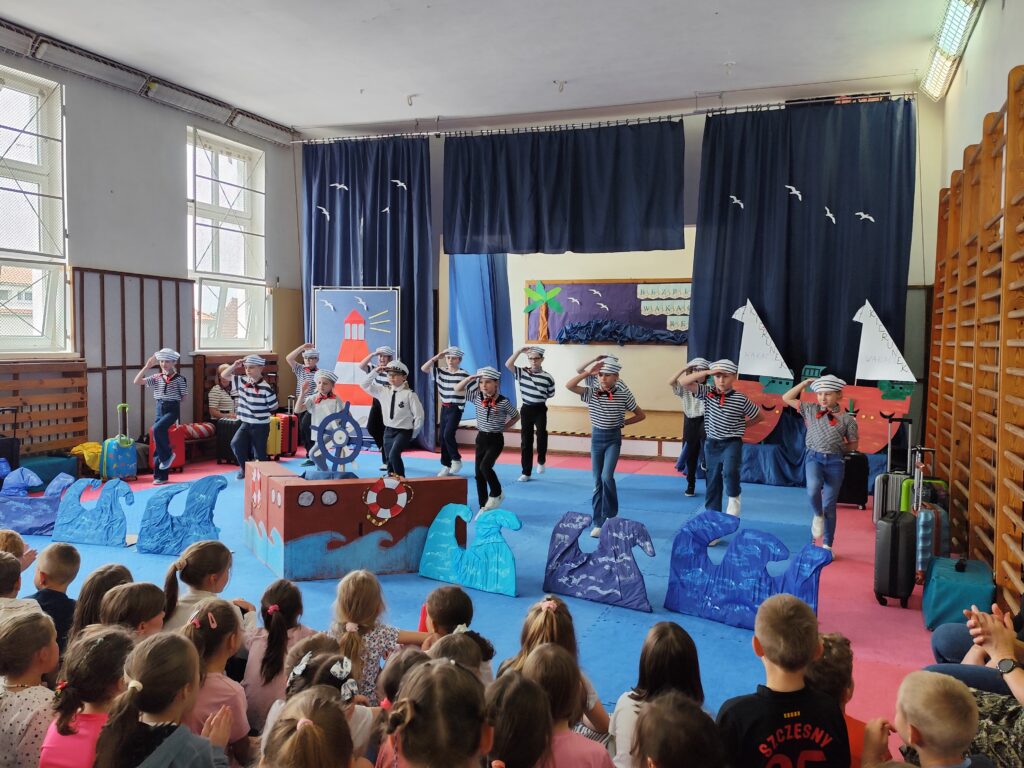 Na zdjęciu widzimy barwne i radosne przedstawienie szkolne. Na pierwszym planie znajduje się grupa uczniów przebranych za marynarzy, w charakterystycznych czapkach i pasiastych koszulkach. Dzieci wykonują układ choreograficzny. W tle widoczna jest scenografia inspirowana motywami morskimi – są fale, ster statku, dekoracje przedstawiające żaglówki i latarnię morską, a także palmę i walizki, nawiązujące do wakacyjnych podróży. Na widowni siedzą uczniowie, z zainteresowaniem i uśmiechem obserwujący występ swoich koleżanek i kolegów.