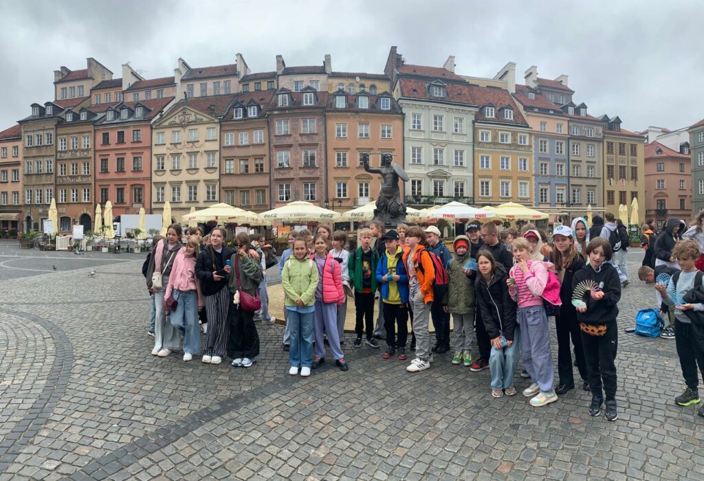Na zdjęciu widać grupę dzieci stojącą na starówce w Warszawie, w pobliżu słynnego pomnika Syrenki Warszawskiej. Dzieci ubrane są w kolorowe kurtki i większość ma plecaki lub torebki. W tle widoczne są charakterystyczne kamienice starego miasta, brukowana nawierzchnia rynku oraz turyści i przechodnie. Atmosfera zdjęcia jest radosna i pełna życia, a dzieci wyglądają na zaciekawione i zaangażowane.