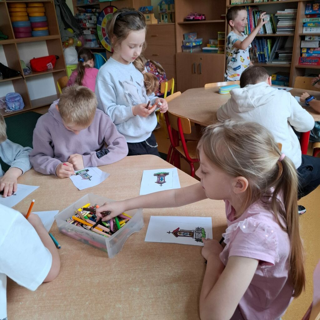 Na zdjęciu widać grupę dzieci pracujących przy stolikach w świetlicy. Dzieci kolorują obrazki przedstawiające warmińskie kapliczki, korzystając z kredek i pisaków. W centrum zdjęcia dziewczynka w szarej bluzie przygląda się swojej pracy, a obok niej chłopcy i dziewczynki skupieni są na rysowaniu. W tle inne dzieci bawią się lub przeglądają książki na regałach. Atmosfera na zdjęciu jest radosna i twórcza, a dzieci zaangażowane są w aktywność plastyczną związaną z poznawaniem kultury Warmii.