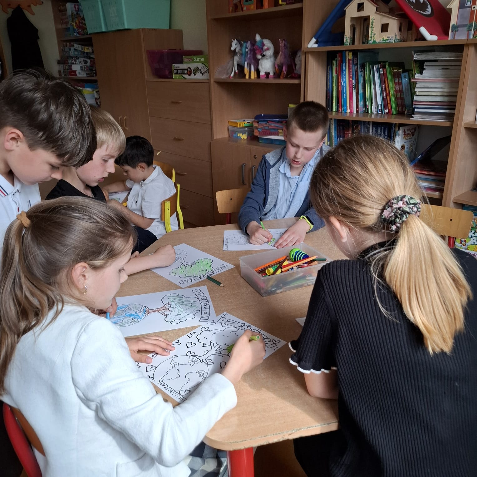 Na zdjęciu widać grupę dzieci siedzących przy stole i kolorujących obrazki. Każde dziecko skupione jest na swojej pracy, używając kredek i flamastrów z plastikowego pojemnika na środku stołu. W tle widać półki z książkami, zabawkami i kolorowymi figurkami, co sugeruje, że zdjęcie zostało wykonane w klasie lub świetlicy szkolnej. Atmosfera wydaje się być spokojna i twórcza, sprzyjająca nauce i zabawie.