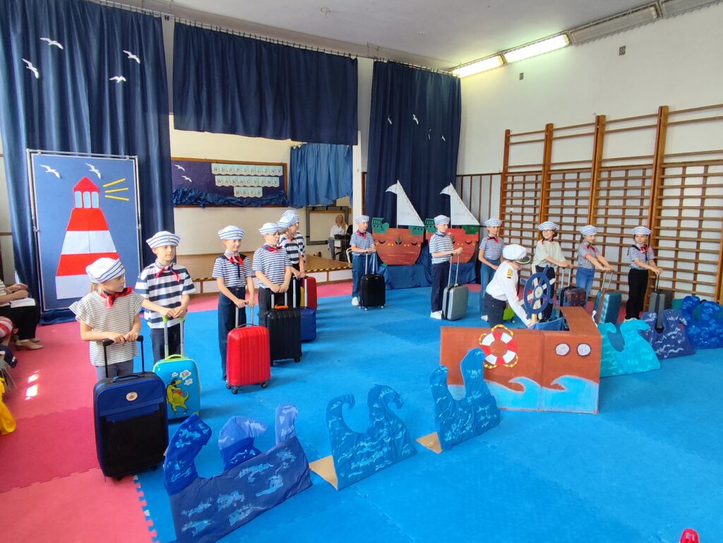 Na zdjęciu widzimy grupę uczniów ubranych w morskie stroje – biało-granatowe koszulki, czapki marynarskie i czerwone apaszki – trzyma kolorowe walizki. Na środku sceny jedno z dzieci, przebrane za kapitana, stoi przy dużym, kartonowym sterze statku. Tło sceny zostało starannie udekorowane. Widzimy niebieskie fale z kartonu, latarnię morską, żaglówki z napisem "Bezpieczne Wakacje", oraz napis „Bezpieczny wakacyjny rejs” umieszczony na tylnej tablicy.