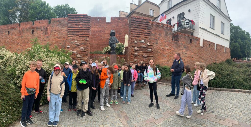 Na zdjęciu widzimy grupę dzieci wraz z przewodnikiem podczas wycieczki szkolnej. Stoją przed Pomnikiem Małego Powstańca w Warszawie, który znajduje się przy murach Starego Miasta. W tle widoczne są czerwone ceglane mury obronne oraz zabytkowe budynki. Dzieci ubrane są na sportowo, a atmosfera wydaje się być pogodna i pełna zainteresowania historią miejsca.
