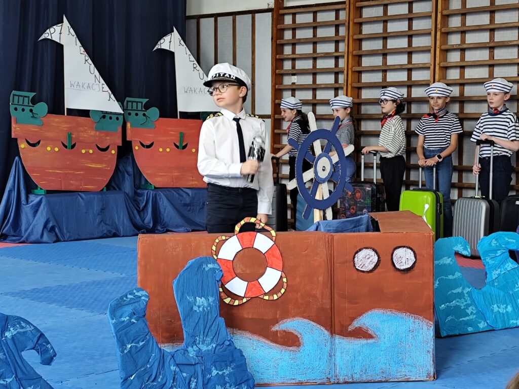 Na zdjęciu widzimy moment ze szkolnego przedstawienia, w którym dzieci wcielają się w załogę statku wyruszającego w wakacyjną podróż. Na pierwszym planie stoi uczeń przebrany za kapitana statku – ma na sobie białą koszulę, ciemne spodnie, krawat i czapkę z napisem „KAPITAN”. Trzyma w dłoni lornetkę i spogląda w dal. Obok niego znajduje się duży, kartonowy model statku z kołem sterowym i malowanymi falami, co nadaje scenie morskiego klimatu. W tle widzimy rząd uczniów przebranych za marynarzy. Każde z dzieci trzyma walizkę, co symbolizuje przygotowanie do wakacyjnej wyprawy. Za nimi umieszczono dekoracje w kształcie statków z żaglami, na których widnieje napis „Kierunek: wakacje”. Całość dopełniają błękitne fale z kartonu i ciemnoniebieskie tło z zawieszonymi mewami, budując atmosferę nadchodzącego lata.
