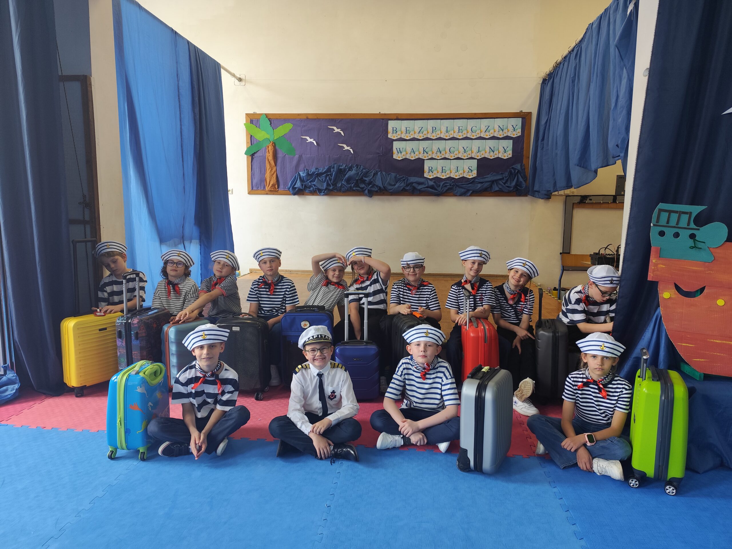 Na zdjęciu widzimy siedzącą na scenie grupę uczniów ubranych w morskie stroje – biało-granatowe koszulki, czapki marynarskie i czerwone apaszki . Obok nich stoją kolorowe walizki. Po prawej stronie widać fragment statku z kartonu na niebieskim tle. Za nimi widzimy tablicę, a na niej palmę, fale morskie, mewy i napis „Bezpieczny wakacyjny rejs”. Wszystko utrzymane jest w morskim klimacie.