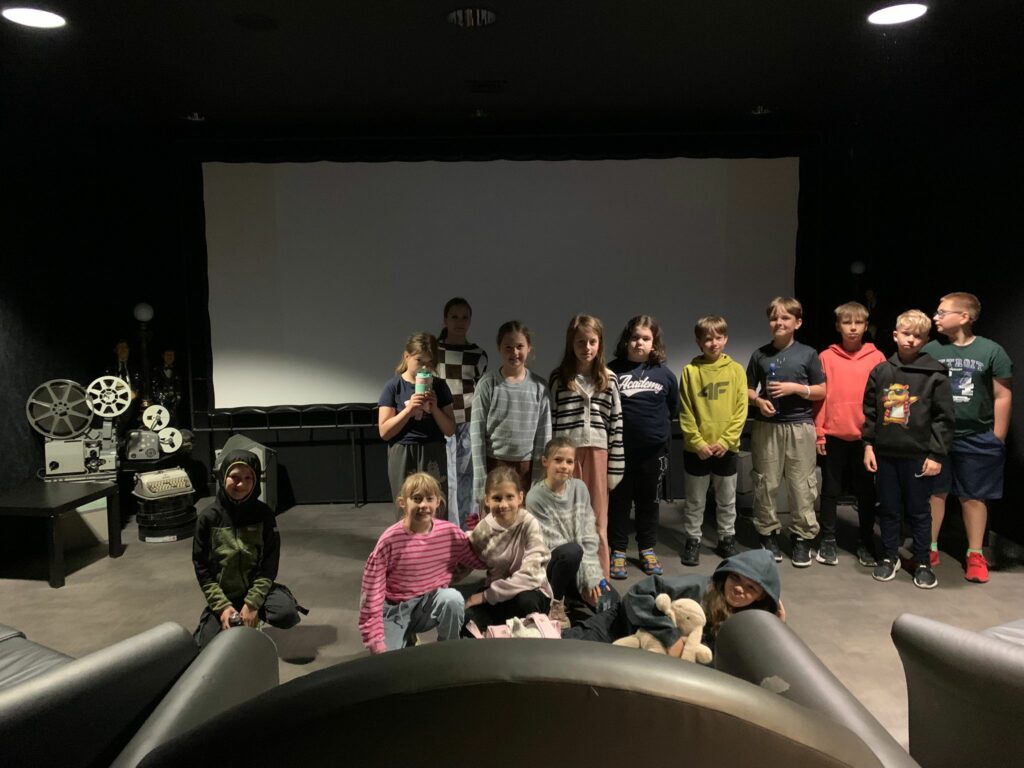 Na zdjęciu widzimy grupę uśmiechniętych dzieci. Zdjęcie zostało zrobione w sali kinowej z czerwonym dywanem i ciemnymi ścianami, na tle dużego, jasnego ekranu projekcyjnego. Część dzieci stoi, niektóre siedzą.
 