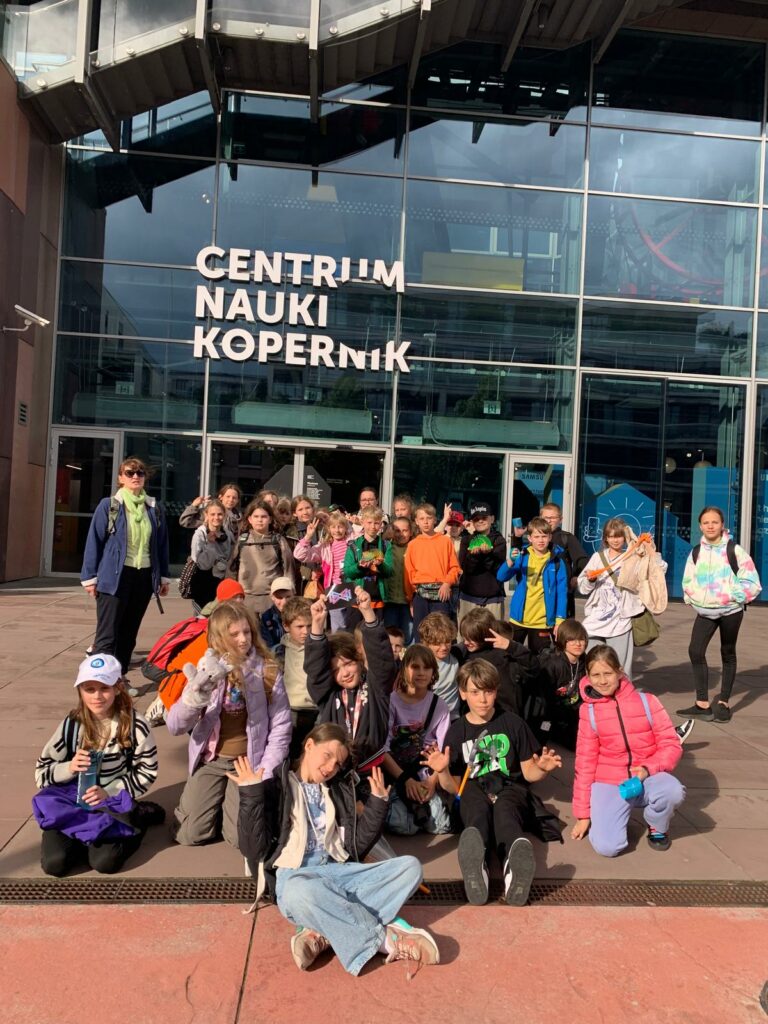 Na zdjęciu widać grupę dzieci wraz z opiekunką stojących przed wejściem do Centrum Nauki Kopernik w Warszawie. Dzieci są uśmiechnięte, niektóre pozują, siedząc na ziemi. Pogoda jest słoneczna, a wszyscy wyglądają na zadowolonych, co sugeruje, że są na szkolnej wycieczce. W tle widoczna jest nowoczesna szklana fasada budynku z dużym napisem „CENTRUM NAUKI KOPERNIK”.