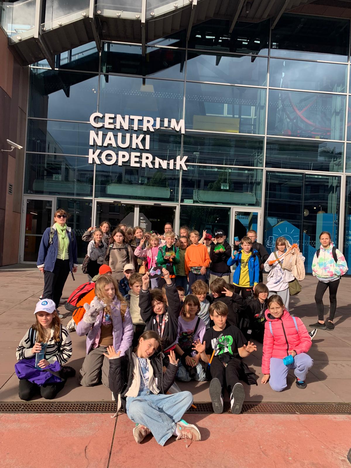 Na zdjęciu widać grupę dzieci wraz z opiekunką stojących przed wejściem do Centrum Nauki Kopernik w Warszawie. Dzieci są uśmiechnięte, niektóre pozują, siedząc na ziemi. Pogoda jest słoneczna, a wszyscy wyglądają na zadowolonych, co sugeruje, że są na szkolnej wycieczce. W tle widoczna jest nowoczesna szklana fasada budynku z dużym napisem „CENTRUM NAUKI KOPERNIK”.