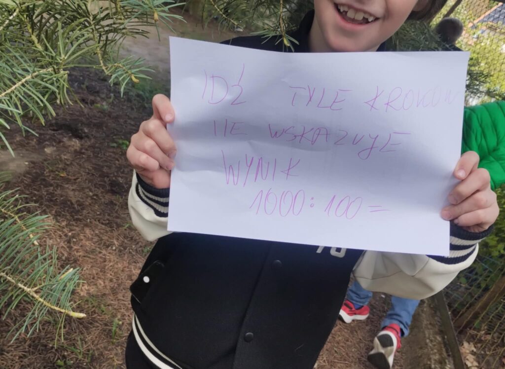 Na zdjęciu widzimy uśmiechnięte dziecko trzymające kartkę z zadaniem matematyczno-ruchowym, które jest częścią gry terenowej. Treść kartki (napisana ręcznie):„Idź tyle kroków ile wskazuje wynik 1000 : 100 =”