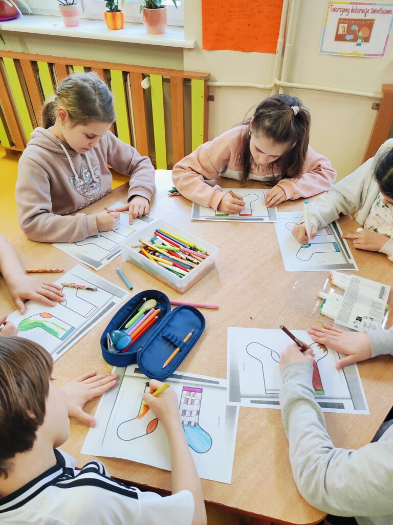 Na zdjęciu widać grupę dzieci siedzących przy stole, którzy są zaangażowani w kolorowanie rysunków. Każde dziecko ma przed sobą kartkę z konturem skarpety, którą koloruje przy użyciu kredek i flamastrów. Na stole znajdują się piórniki i przybory do pisania. W tle zdjęcia widać doniczki z roślinami ustawione na białym parapecie oraz kolorowe plakaty.