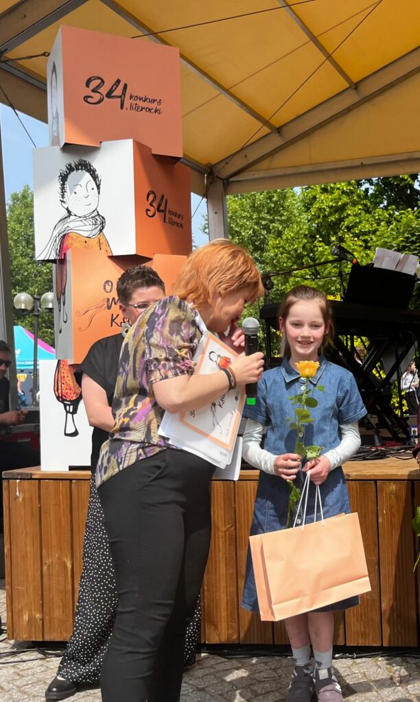 Na zdjęciu widzimy kobietę o kasztanowych włosach, która wręcza dziewczynce nagrodę żółtą różę, dyplom i papierową torebkę. Dziewczynka jest uśmiechnięta, ubrana w sukienkę dżinsową. W tle widać scenę, na scenie stoją instrumenty muzyczne oraz dekoracja 34 konkursu literackiego