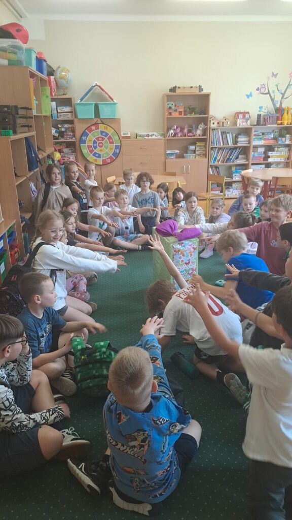 Na zdjęciu widać dzieci siedzące w kole w klasie, z wyciągniętymi rękami w stronę dużego, kolorowo zapakowanego prezentu ustawionego na środku sali. Dzieci są uśmiechnięte – czekają na moment otwarcia niespodzianki. W tle widać regały z książkami, grami i zabawkami. Zdjęcie wykonano w świetlicy szkolnej.