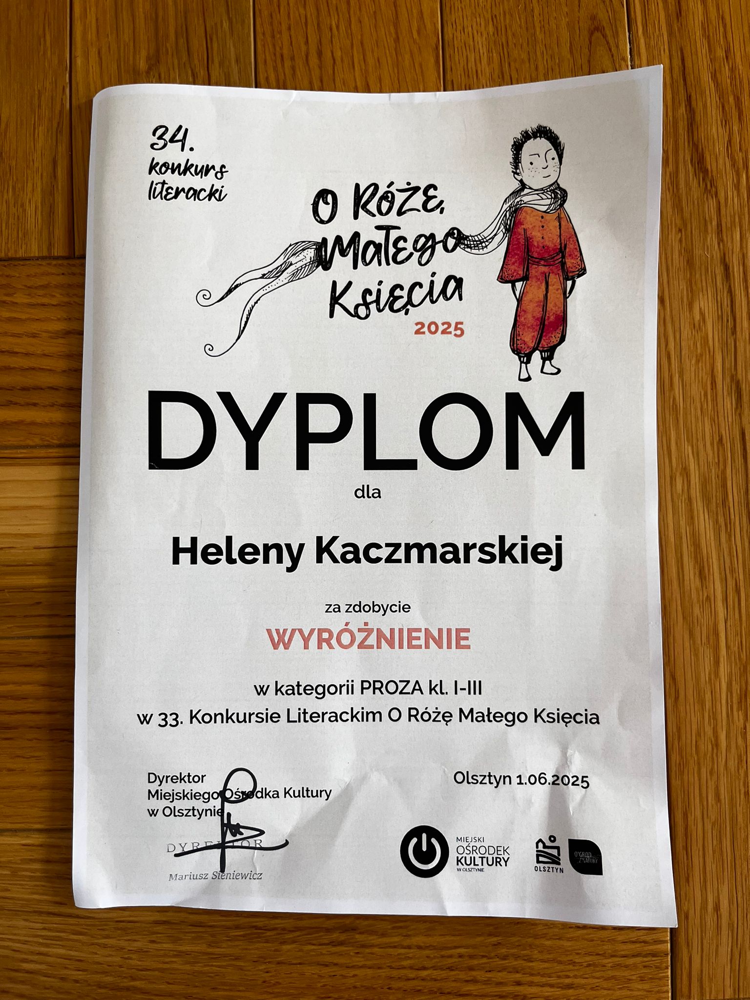 Na zdjęciu widać dyplom dla Heleny Kaczmarskiej za zdobycie wyróżnienia w kategorii klas I-III w 34 Konkursie Literackim O Różę Małego Księcia. Na górze po lewej stronie 34. Koknkurs Literacki. Po prawej stronie na górze napis O Różę Małego Księcia 2025 i obrazek Ksiecia w czerwonym kombinezonie i szaliku białym. Na dole po lewej stronie pieczątka Dyrektora Ośrodka Kultury w Olsztynie - Mariusz Stenewicz. Po prawej stronie na dole Olsztyn 1.06.2025.