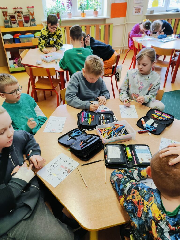 Kilka dzieci siedzi przy okrągłym stole i rysuje na kartkach z kształtami puzzli. Na stole widać liczne piórniki, kredki, nożyczki i inne przybory plastyczne. Jedno z dzieci ma rękę w temblaku, ale mimo to aktywnie uczestniczy w zajęciach. Dzieci współpracują i rozmawiają, co świadczy o wspólnej zabawie i kreatywnym działaniu.