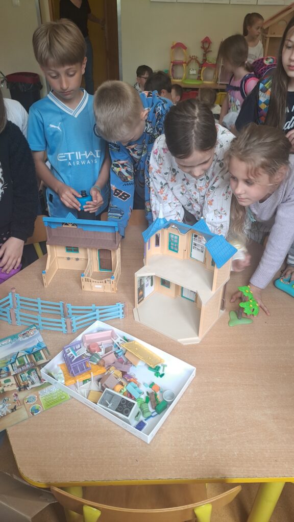 Na zdjęciu są dzieci pochylone nad stołem, na którym znajduje się domek Playmobil do składania wraz z akcesoriami – mebelkami i figurkami. Dzieci oglądają elementy zestawu.