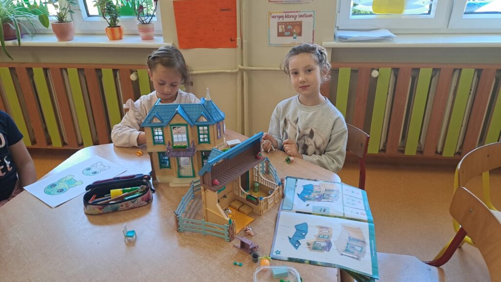 Na zdjęciu widać dwie dziewczynki siedzące przy stoliku, na którym stoi domek Playmobile, leży piórnik, instrukcja składania domku Playmobile oraz kartka z namalowanymi żółwiami.