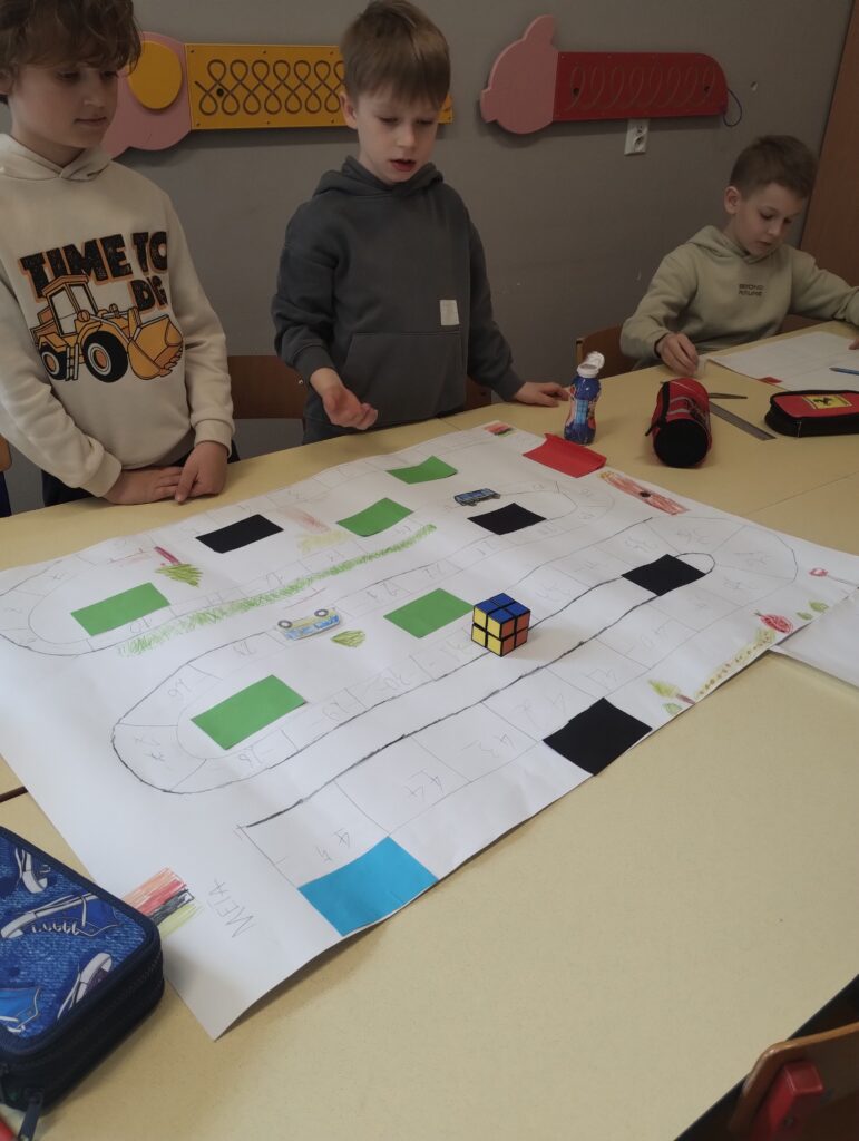 Na zdjęciu widzimy trójkę chłopców siedzących i stojących przy szkolnej ławce, nad którą rozłożona jest własnoręcznie wykonana gra planszowa. Plansza została wykonana na dużym arkuszu papieru i przypomina tor gry z ponumerowanymi polami. Pojawiają się też kolorowe kwadraty (zielone, czarne, niebieskie i czerwone), które mogą mieć specjalne znaczenie w grze (np. bonusy, przeszkody, zadania). Na planszy znajduje się kostka Rubika – używana jako pionek lub element gry. Jeden z chłopców gestykuluje, jakby tłumaczył zasady lub był właśnie w trakcie ruchu. Pozostali uczestnicy są skupieni i zainteresowani rozgrywką. W tle widać typową klasę szkolną z elementami dekoracyjnymi na ścianie (m.in. zabawkowe narzędzia lub kolorowe tablice manipulacyjne).