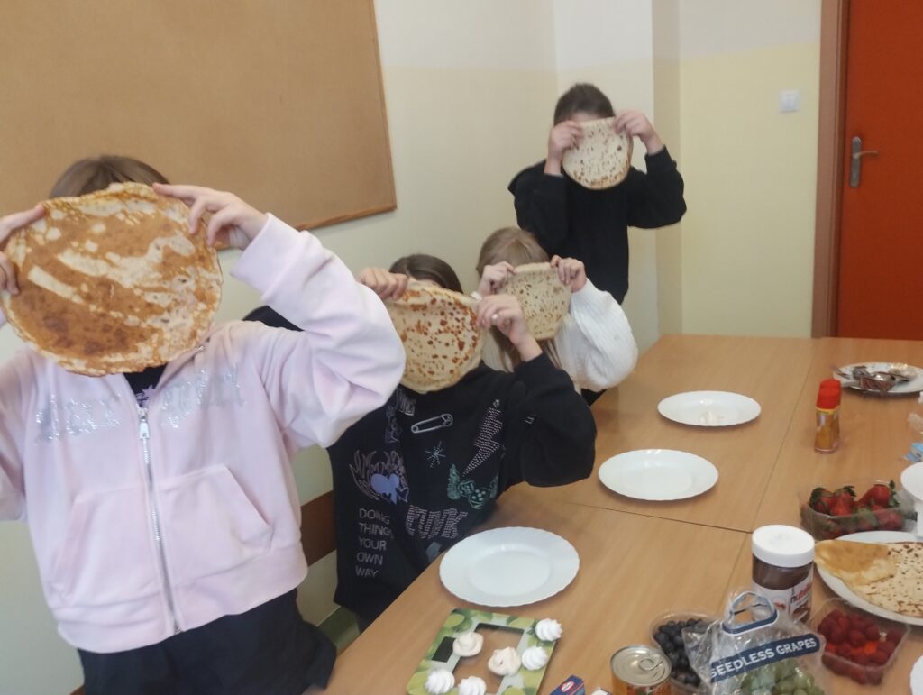 Na zdjęciu widzimy grupę dzieci uczestniczących w kulinarnych zajęciach w klasie. Dzieci siedzą lub stoją przy stole i trzymają przed twarzami naleśniki, zasłaniając nimi swoje twarze – wygląda to zabawnie i świadczy o dobrej zabawie.Na stole znajdują się produkty spożywcze: owoce (truskawki, winogrona, maliny), Nutella, bita śmietana, bezy, krem w sprayu, puste talerze – prawdopodobnie przygotowane na samodzielne tworzenie słodkich naleśników. W tle widać salę z tablicą korkową na ścianie i drzwiami. Atmosfera jest wesoła i swobodna, a uczniowie mają okazję nie tylko do nauki przez działanie, ale też do integracji i zabawy.