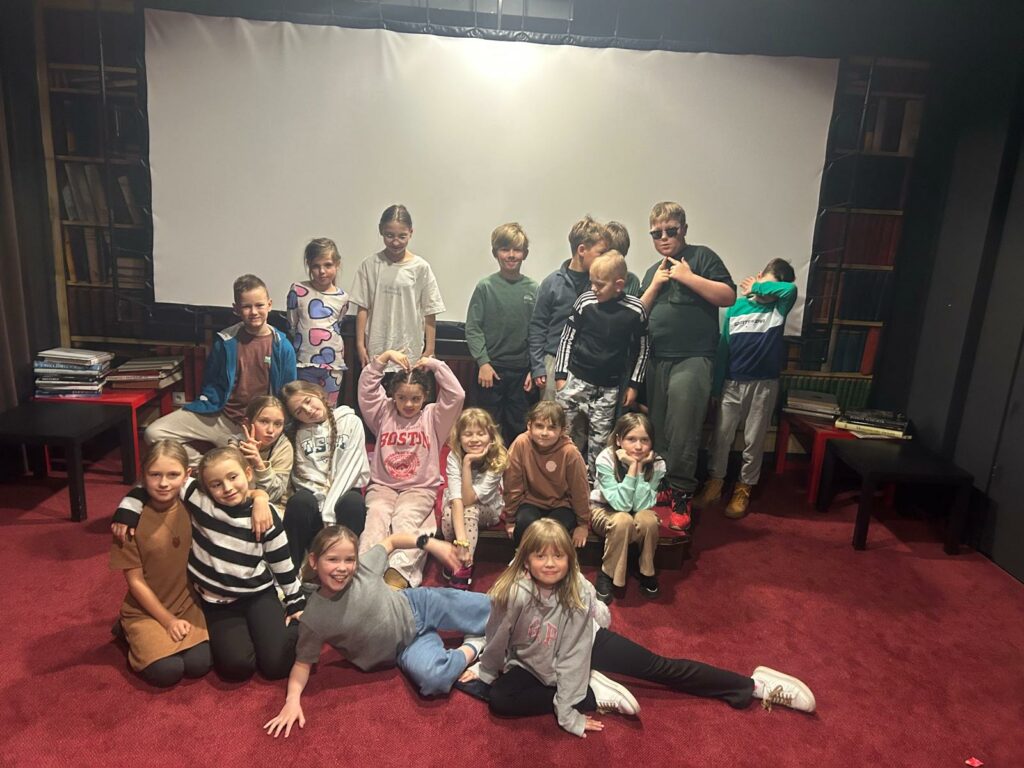 Na zdjęciu widzimy grupę uśmiechniętych dzieci w różnym wieku, prawdopodobnie z jednej klasy lub grupy zajęciowej. Zdjęcie zostało zrobione w sali kinowej z czerwonym dywanem i ciemnymi ścianami, na tle dużego, jasnego ekranu projekcyjnego. W tle widać także regały z książkami – miejsce ma kameralny, przytulny charakter. Dzieci pozują w swobodny, radosny sposób – część siedzi, część klęczy, inne stoją, niektóre wykonują zabawne pozy i gesty, co nadaje zdjęciu bardzo ciepły i naturalny klimat. Wszyscy wyglądają na zrelaksowanych i zadowolonych.