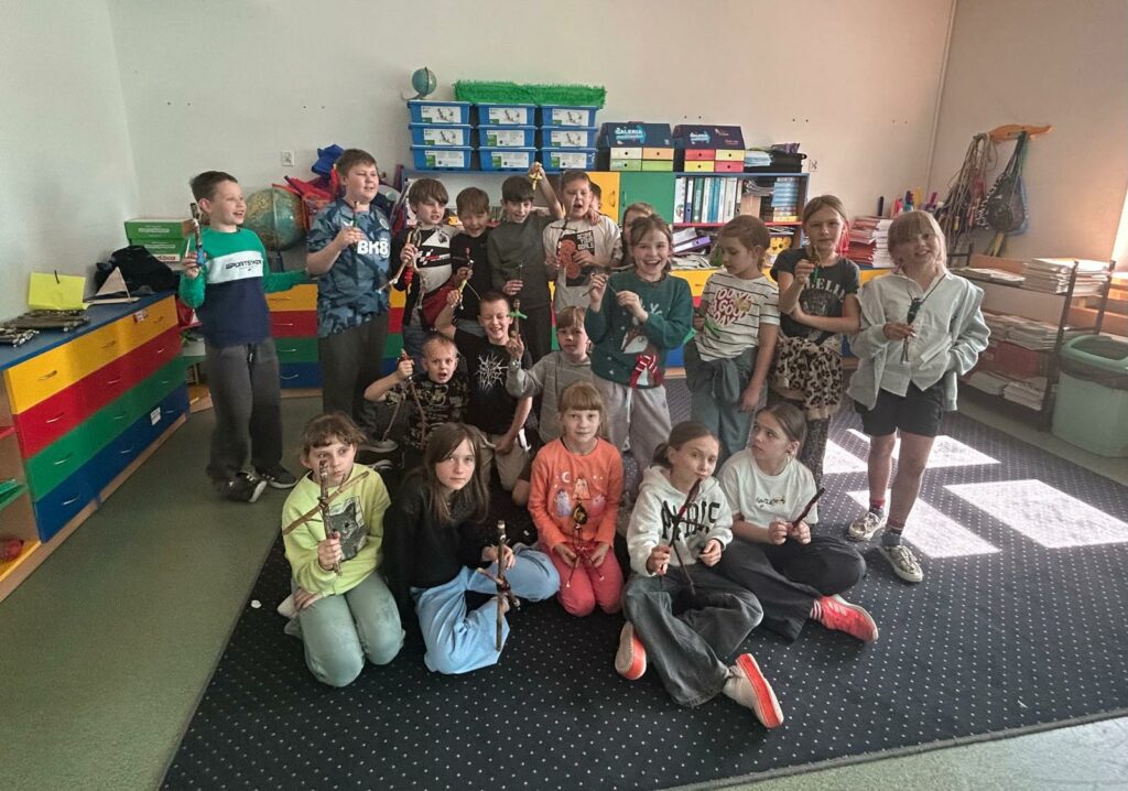 Na zdjęciu widzimy grupę dzieci w sali lekcyjnej, które prezentują własnoręcznie wykonane prace plastyczne – ludziki lub figurki z patyczków. Atmosfera jest radosna i pełna energii, dzieci uśmiechają się i z entuzjazmem pozują do wspólnej fotografii. W tle kolorowe szafki i półki z materiałami dydaktycznymi – typowe dla klas wczesnoszkolnych. Widać też przestrzeń sprzyjającą twórczej pracy.