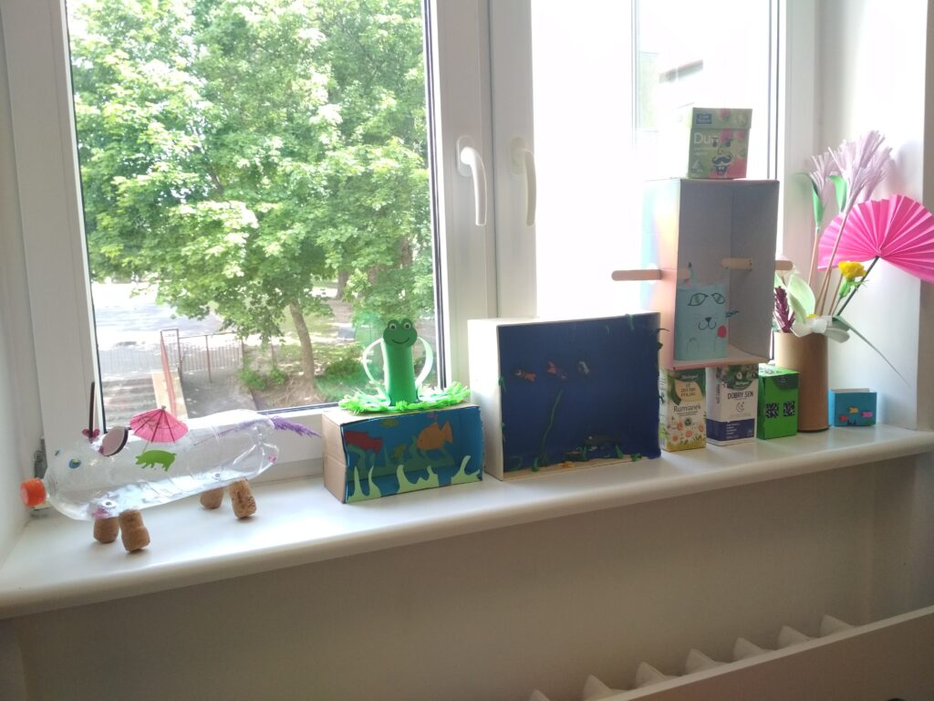 Przedstawia formy przestrzenne, w których wykorzystano surowce wtórne. Ustawione są one w formie wystawki, od prawej strony: akwarium, wazon z kwiatami, domek, robot, kot, 2 akwaria, żaba.