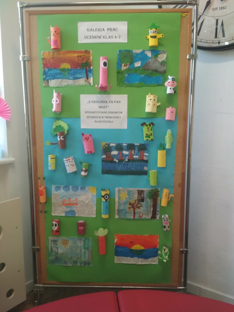Przedstawia stojącą tablicę ścienną, na której umieszczony jest na górze napis- galeria prac uczniów klas 4-7 .Niżej znajduje się napis-,,Z ekologią za pan brat”. Wykorzystanie surowców wtórnych w twórczości plastycznej , pod nimi znajdują się prace plastyczne przedstawiające krajobrazy, wykonane techniką kolażu. Dookoła prac i napisów rozmieszczone są formy przestrzenne wykonane z rolek od papieru przedstawiające między innymi: kotka, pieska, świnkę, drzewo, ośmiornicę, krówkę, marchewkę ,ananasa.