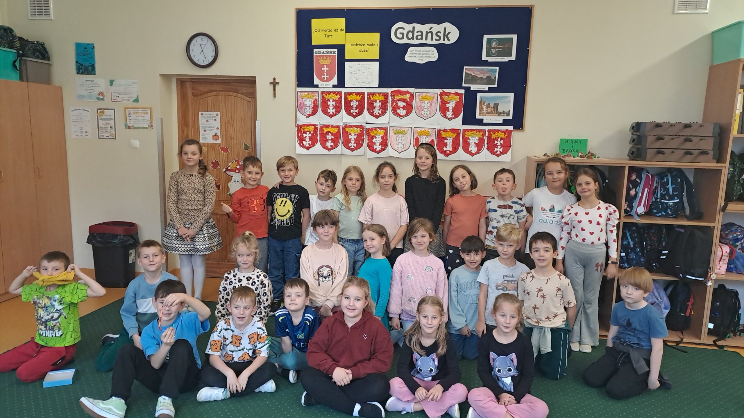 Na zdjęciu widać grupę uśmiechniętych dzieci stojących i siedzących w sali przed tablicą z napisem „Gdańsk” oraz pracami plastycznymi przedstawiającymi herby miasta wyklejone bibułą. W tle znajdują się także zdjęcia zabytków Gdańska i mapka Polski, a dzieci pozują wspólnie do pamiątkowej fotografii.