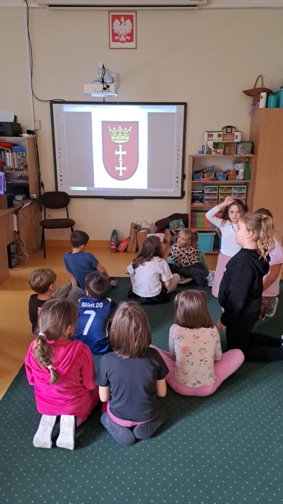 Na zdjęciu widać grupę dzieci siedzących na dywanie w sali , które oglądają na tablicy multimedialnej obraz przedstawiający herb Gdańska – czerwone tło z dwoma białymi krzyżami i złotą koroną. Nad tablicą wisi godło Polski, a w tle stoją półki z książkami i pomocami do zajęć.