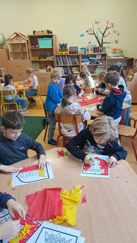 Na zdjęciu widać grupę dzieci siedzących przy stolikach. Dzieci pochylone są nad kartami pracy, na których widnieje herb Gdańska. Na stolikach znajduje się czerwona i żółta bibuła oraz klej. W tle widać półki z książkami i grami planszowymi.