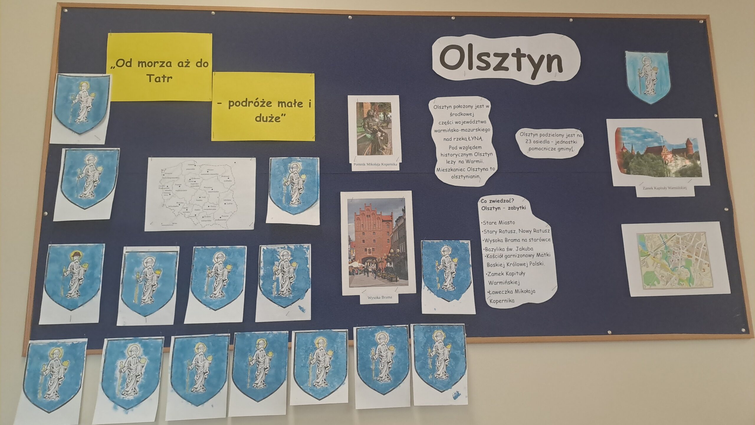 Na zdjęciu widać zieloną tablicę na której jest duży napis Olsztyn, są także informacje o mieście Olsztynie, kolorowe herby Olsztyna, zdjęcia zabytków Olsztyna.