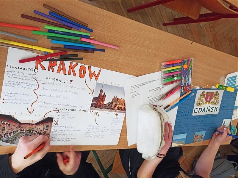 Na zdjęciu widać fragment szkolnej klasy oraz ławkę, przy której uczniowie pracują nad projektem. Na blacie leżą kolorowe flamastry, długopisy, klej w sztyfcie i piórnik. W centrum znajduje się duży plakat z napisem „KRAKÓW”, ozdobiony zdjęciami oraz ręcznie zapisanymi informacjami i ciekawostkami o mieście. Jedna osoba właśnie dorysowuje elementy czerwonym pisakiem. Obok widać drugi plakat dotyczący Gdańska – niebieską kartkę z herbem miasta i notatkami. Uczeń trzyma długopis i dopisuje kolejne informacje. Na ławce leżą też wydrukowane materiały oraz fotografie zabytków, które są przyklejane do projektów.