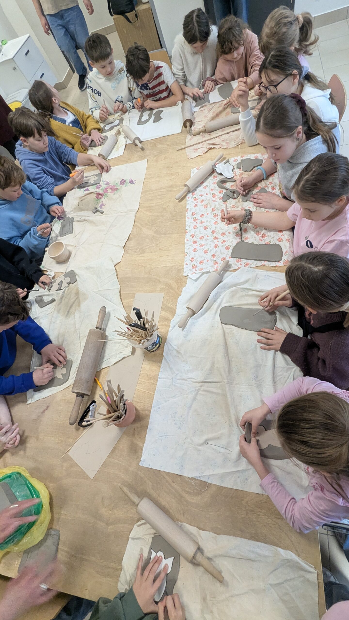To ujęcie pokazuje pracę przy dużym stole warsztatowym. Wokół stołu siedzi i stoi liczna grupa dzieci. Każde dziecko ma przed sobą kawałek gliny lub masy plastycznej. Widać wałki, patyczki, drewniane narzędzia i szablony do formowania. Dzieci wycinają, wygładzają i zdobią swoje prace. Na stole leżą ściereczki i podkładki, które zabezpieczają miejsce pracy. Zdjęcie dobrze oddaje skupienie, współpracę i twórcze zaangażowanie uczestników.