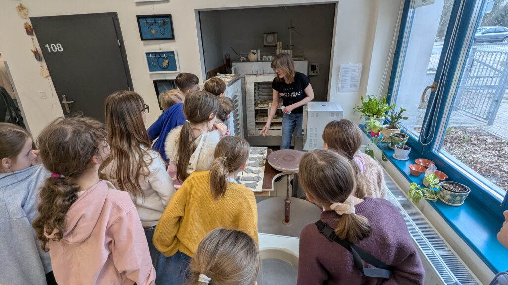 Na tym zdjęciu grupa dzieci stoi w pracowni i słucha prowadzącej, która pokazuje im coś przy piecu ceramicznym lub urządzeniu do wypalania. Dzieci są ustawione półkolem i obserwują demonstrację. Prowadząca wskazuje na elementy znajdujące się przy piecu. Na blacie obok widać ułożone gliniane prace, prawdopodobnie przygotowane do kolejnego etapu. W pomieszczeniu jest dużo światła dziennego, a na parapecie stoją rośliny doniczkowe, co ociepla przestrzeń.