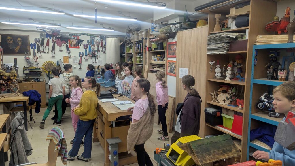 To szerszy widok na całą pracownię artystyczną. Dzieci stoją i rozglądają się po wnętrzu. Pomieszczenie jest pełne rękodzieła, figurek, masek, ozdób, narzędzi i gotowych prac. Na ścianach i półkach widać wiele barwnych, fantazyjnych obiektów artystycznych. W centrum znajduje się stół roboczy, a wokół niego wyposażenie typowe dla pracowni plastycznej lub technicznej. Zdjęcie pokazuje miejsce jako inspirującą, kreatywną przestrzeń, która pobudza wyobraźnię dzieci.