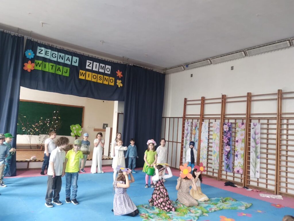 Zdjęcie przedstawia grupę dzieci siedzących i stojących na szkolnej sali gimnastycznej przed sceną udekorowaną na powitanie wiosny. W tle widać kurtynę z napisem „Żegnaj zimo, witaj wiosno” oraz proste dekoracje z kwiatami i gałązkami. Dzieci są ubrane w jasne, kolorowe stroje. Są w trakcie przedstawienia.