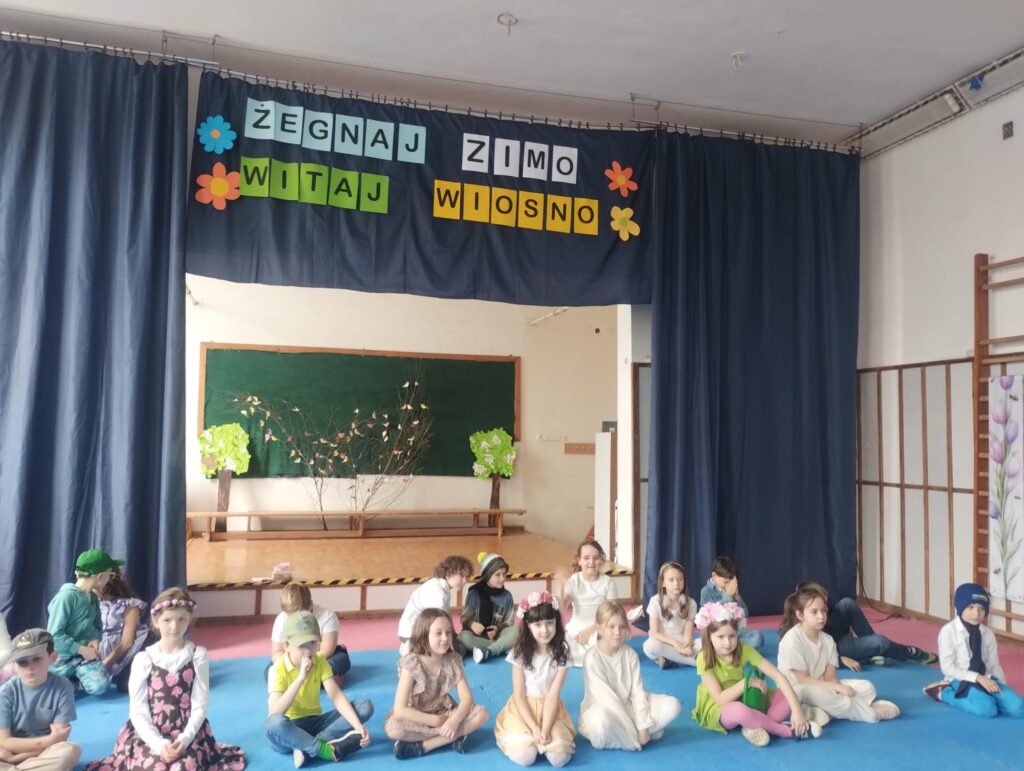 Zdjęcie przedstawia grupę dzieci siedzących w sali szkolnej przed sceną udekorowaną na powitanie wiosny. W tle widać kurtynę z napisem „Żegnaj zimo, witaj wiosno” oraz dekoracje z kwiatami i gałązkami. Dzieci są ubrane w jasne, kolorowe stroje i przygotowują się do występu.