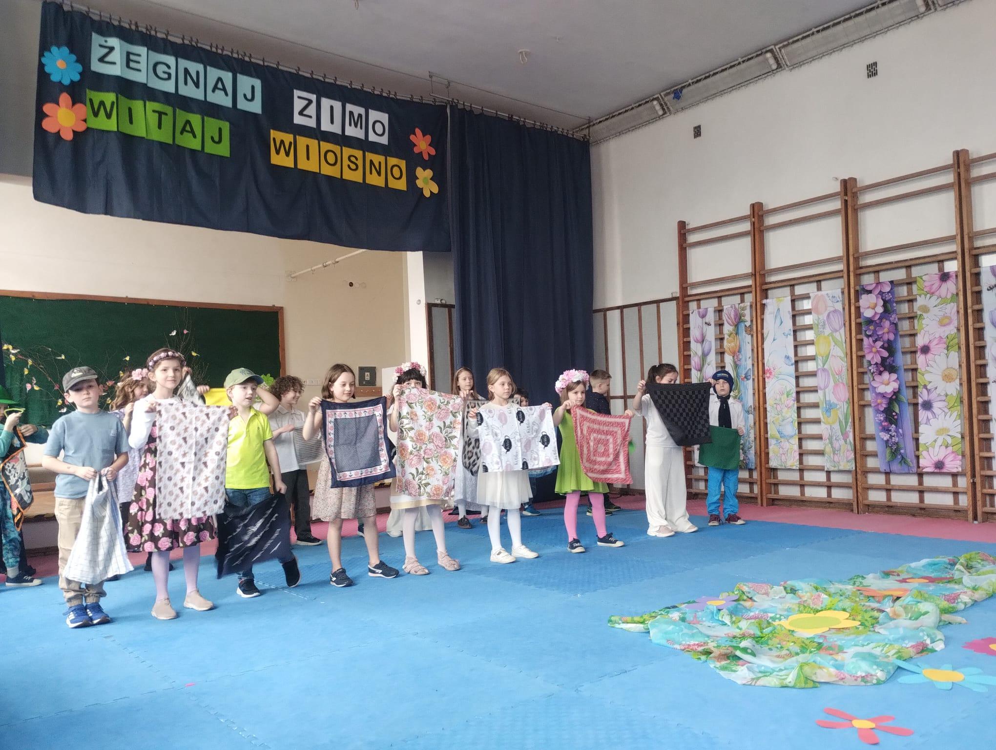 Grupa dzieci stoi w sali gimnastycznej na niebieskiej macie i prezentuje kolorowe chustki podczas występu. W tle widać dekorację z napisem „Żegnaj zimo, witaj wiosno” oraz wiosenne ozdoby z kwiatami.
